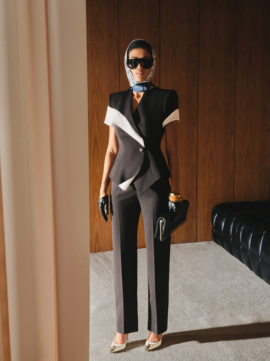 Woman in a stylish outfit with a black and white jacket and gray pants, standing in a room with wooden paneling.