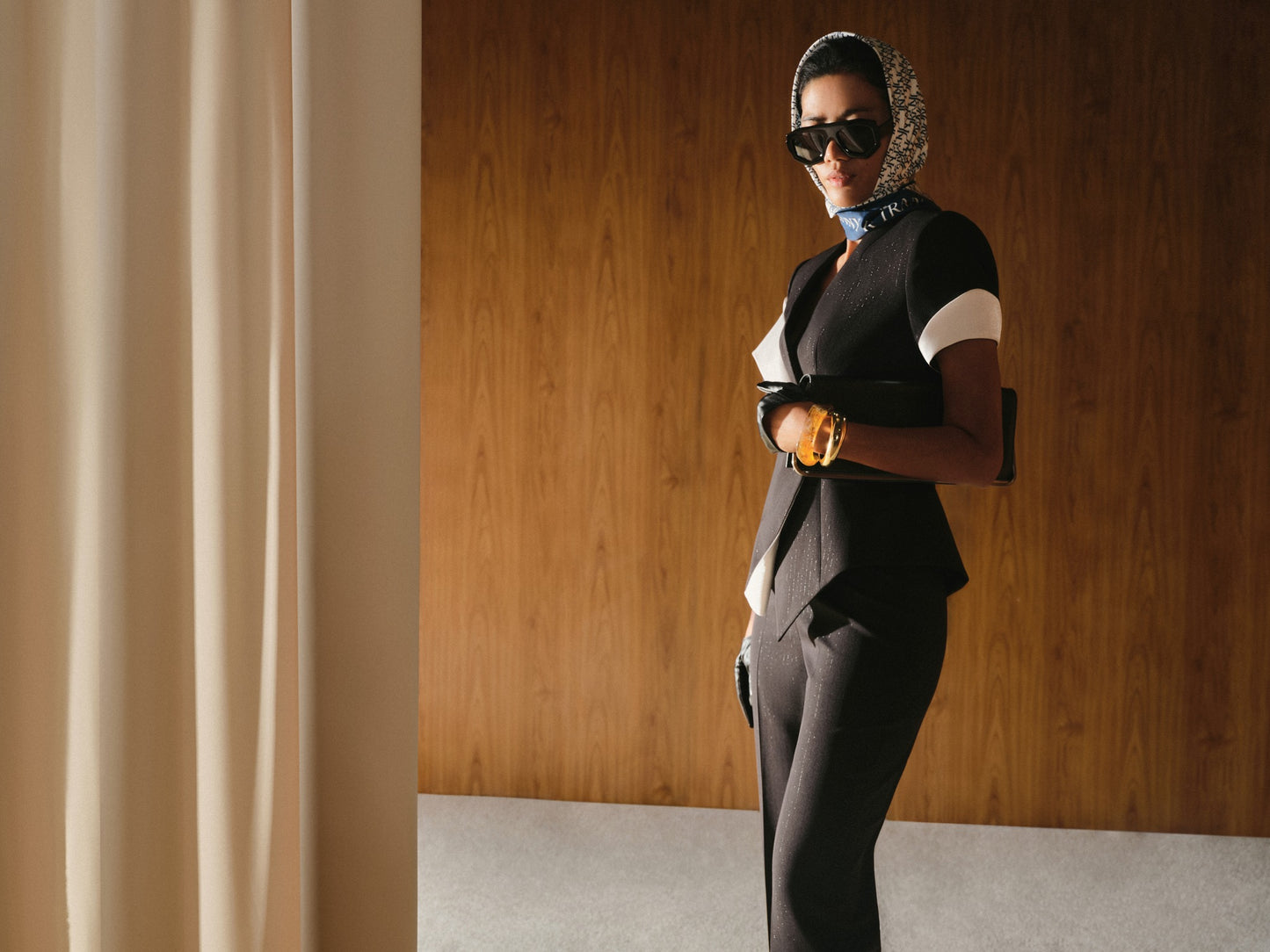 Woman in a stylish outfit with sunglasses and a headscarf against a wooden wall.