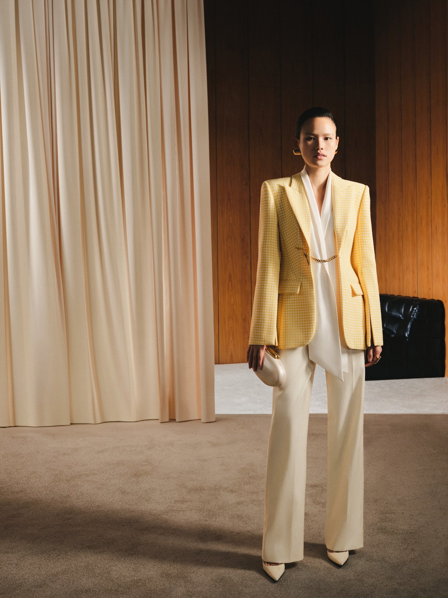 Woman in a yellow blazer and white pants standing in a room with beige curtains and a black sofa.