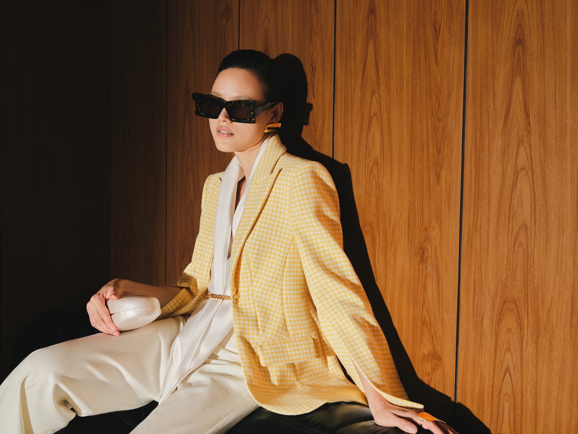 Woman in a yellow checkered blazer and white pants sitting against a wooden wall.