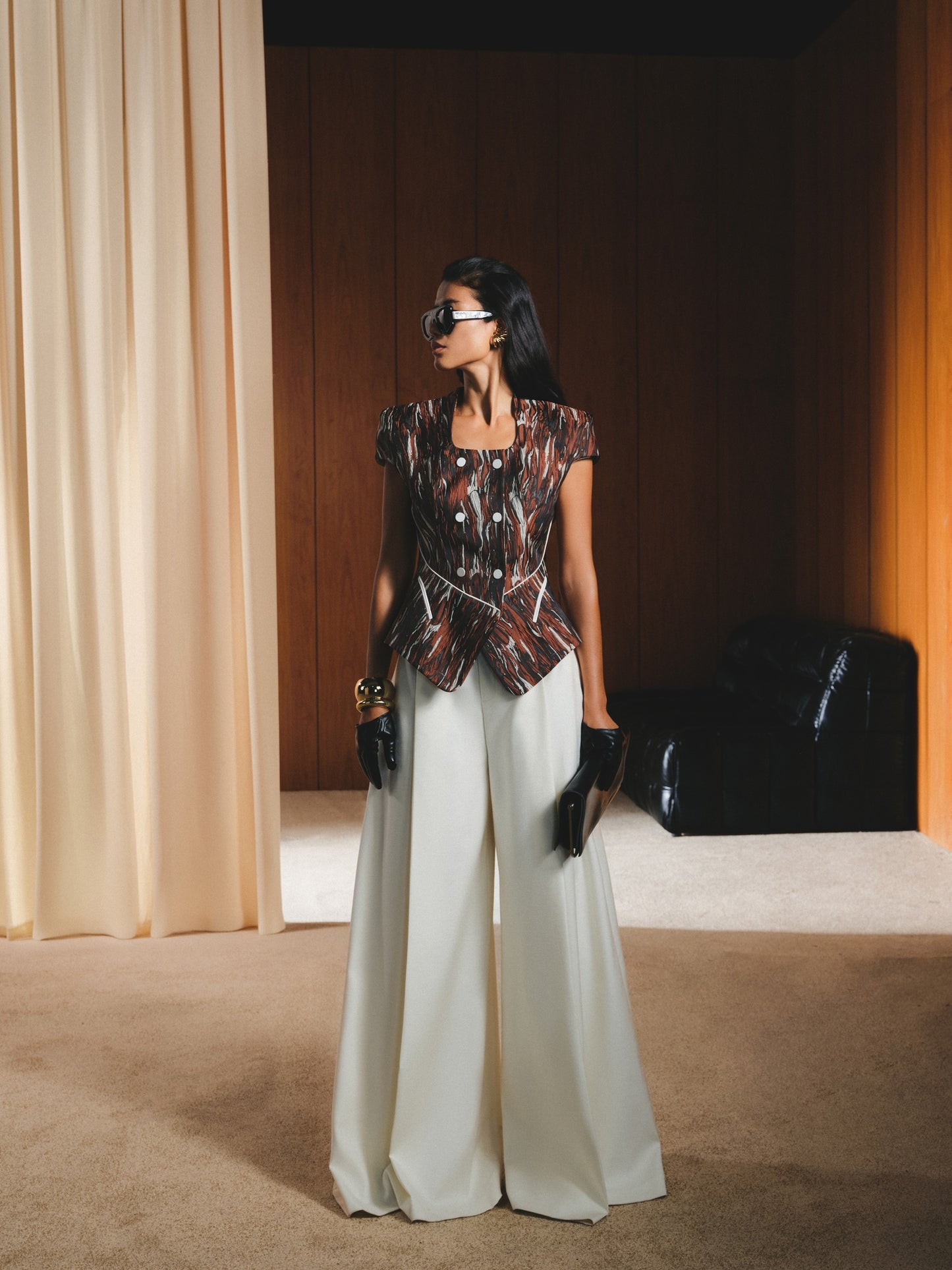 Woman in a patterned top and white pants standing in a room with beige curtains and a black bag.