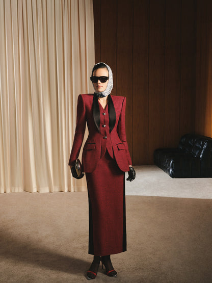 Woman in a red suit standing in a room with beige curtains and a black leather couch.