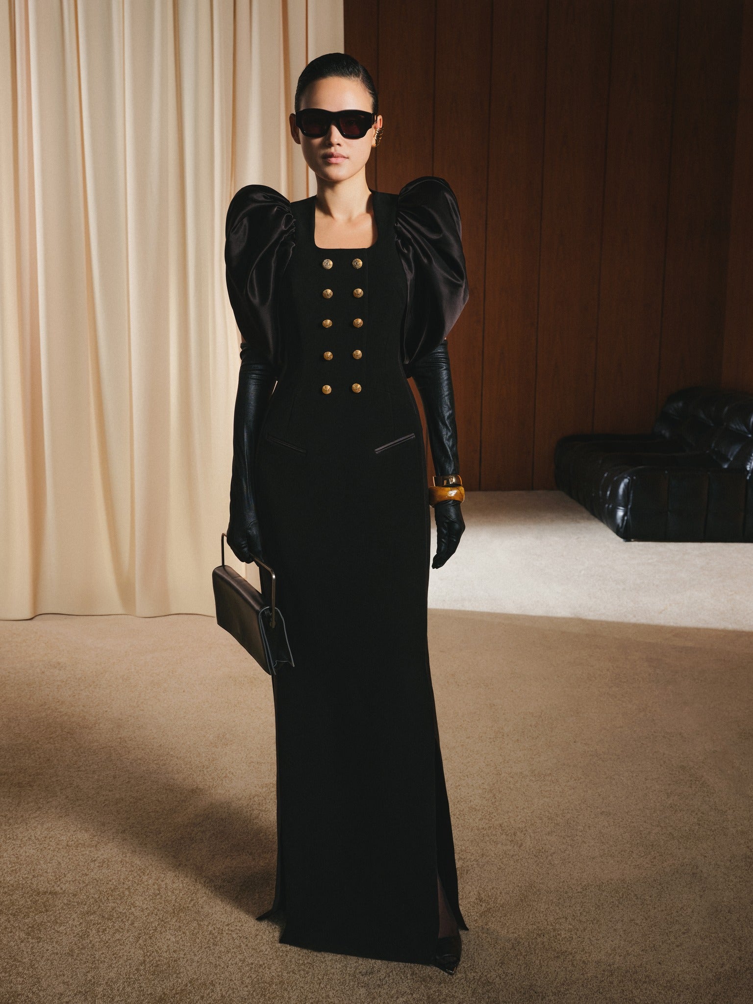 Woman in a black dress with gold buttons standing in a room with beige curtains and a black sofa.