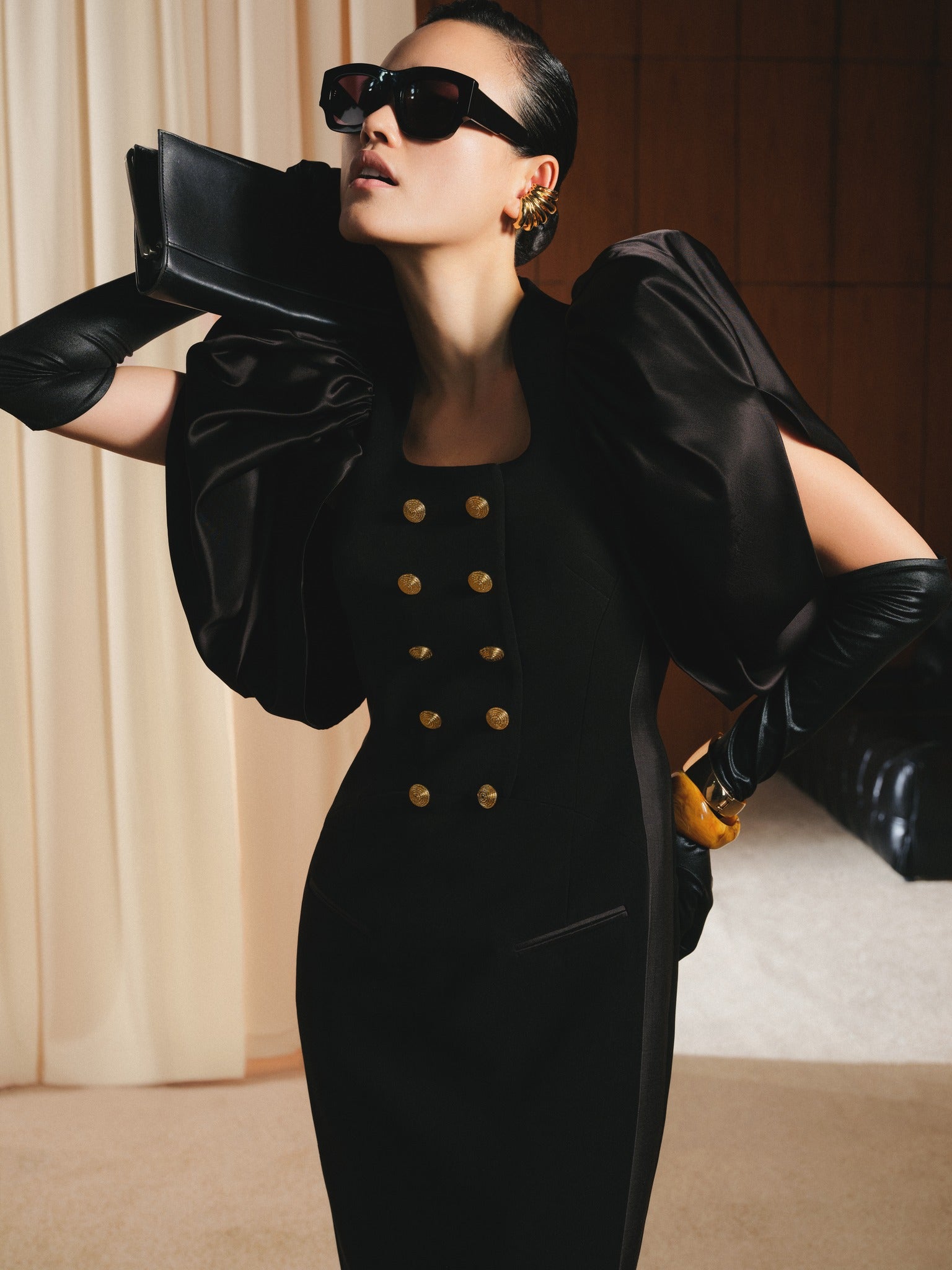 Woman in a black outfit with gold buttons, holding a black clutch, in an indoor setting.