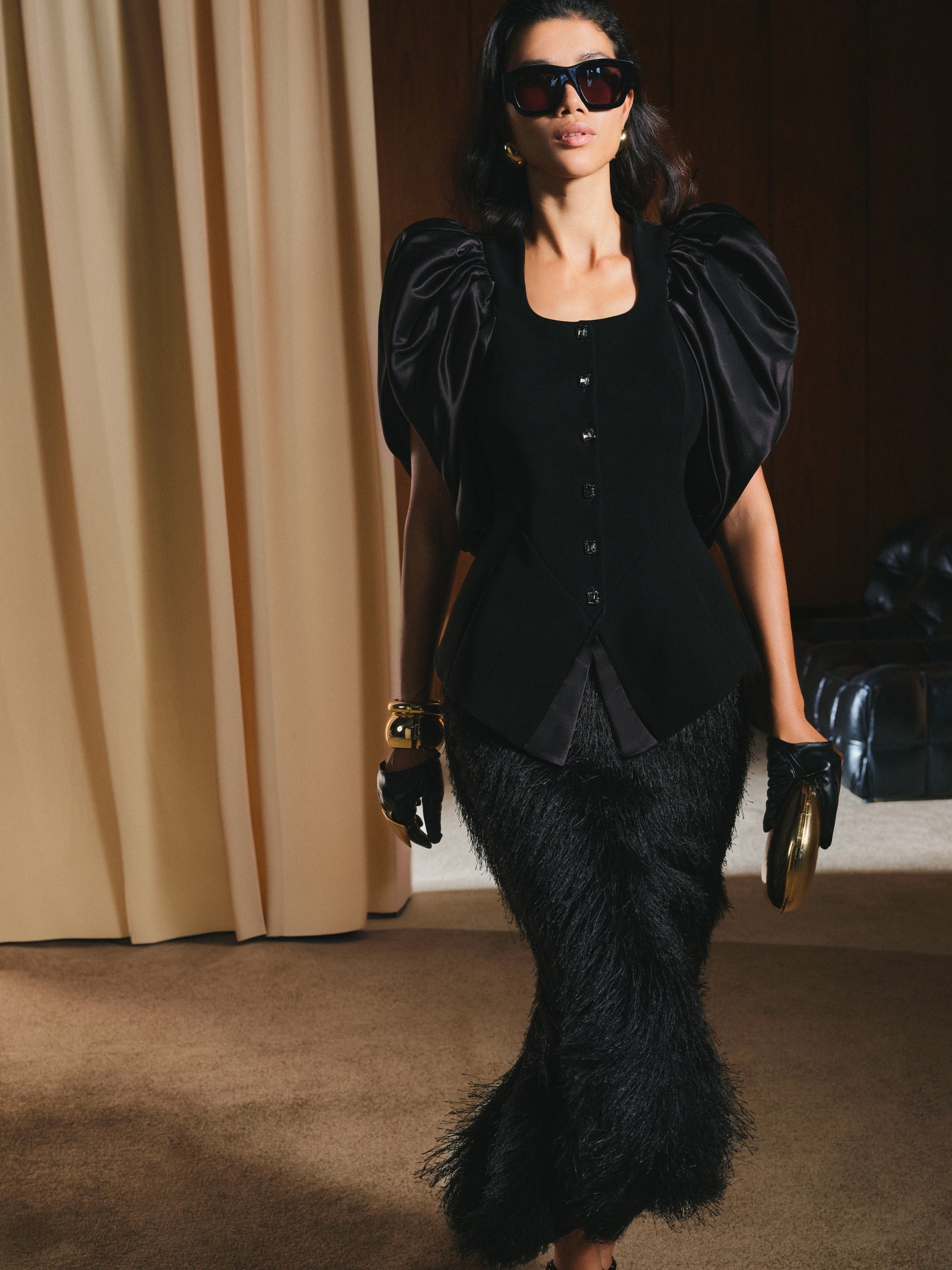 Model wearing a black outfit with puffed sleeves and feathered skirt in a room with beige curtains.