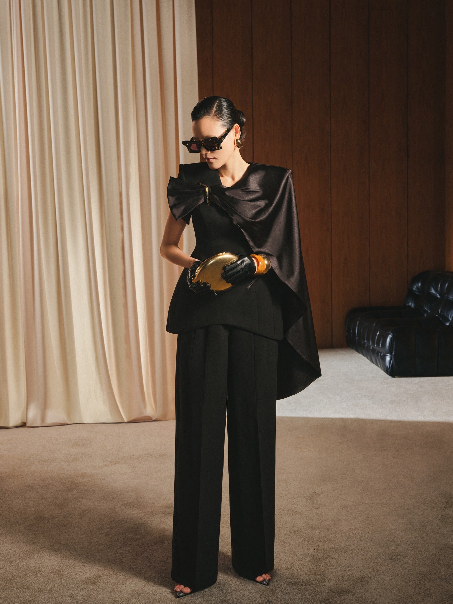 Woman in a black outfit with a cape holding a gold clutch in an elegant room.