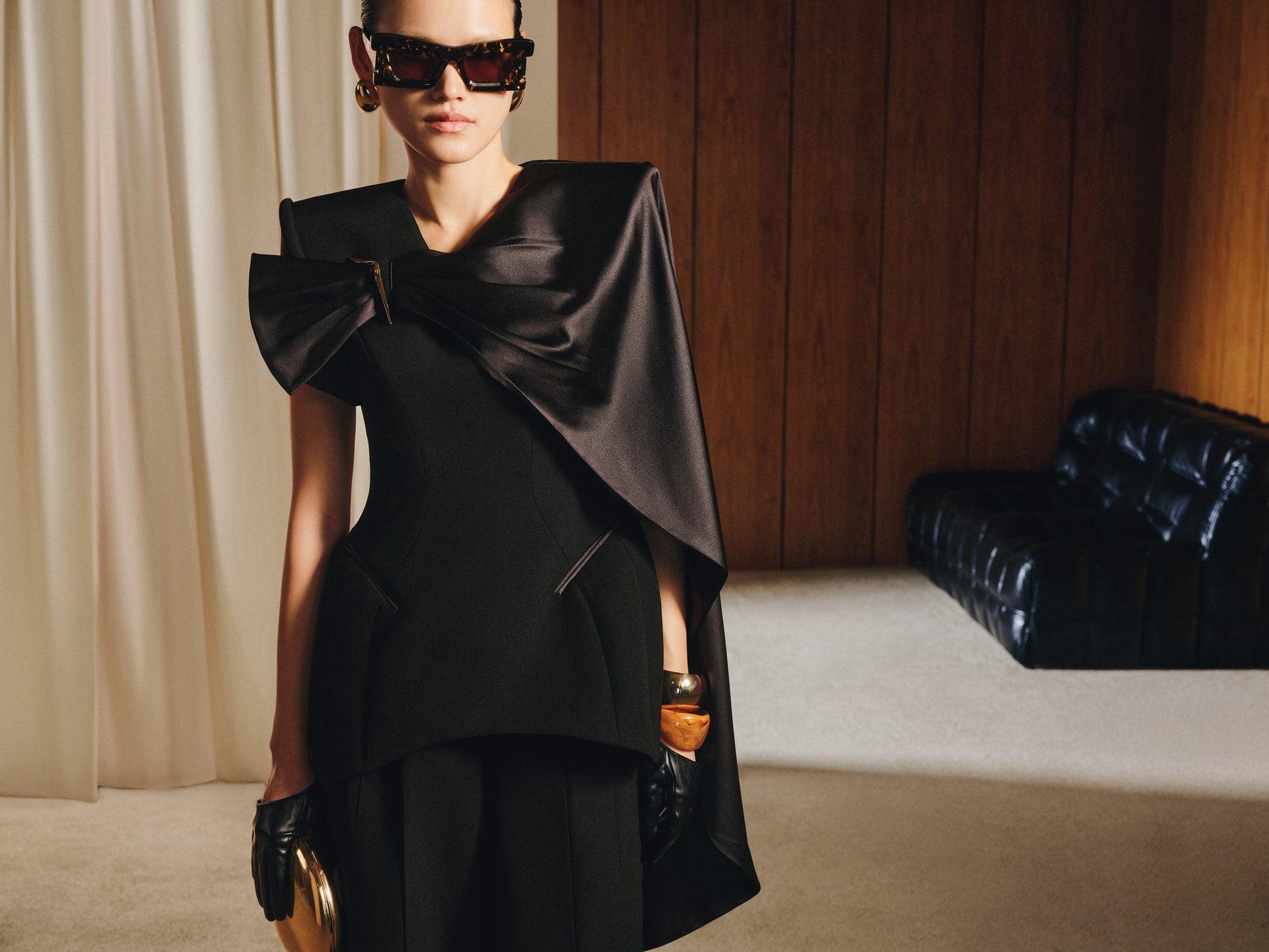 Woman wearing a black dress with a large bow in a room with wooden paneling and a black leather couch.