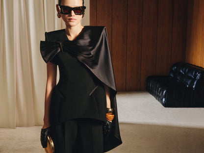 Woman wearing a black dress with a large bow in a room with wooden paneling and a black leather couch.