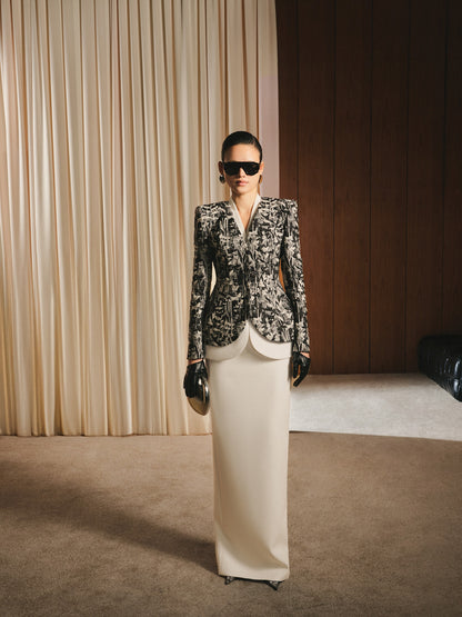 Woman in a patterned blazer and white skirt standing in a room with beige curtains and wooden wall.