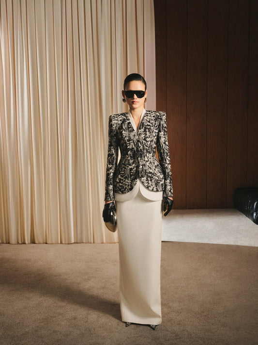 Woman in a patterned blazer and white skirt standing in a room with beige curtains and wooden wall.