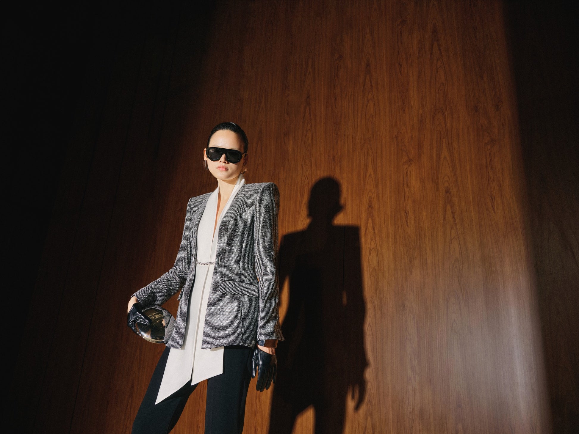 Person wearing sunglasses and a gray blazer against a wooden wall