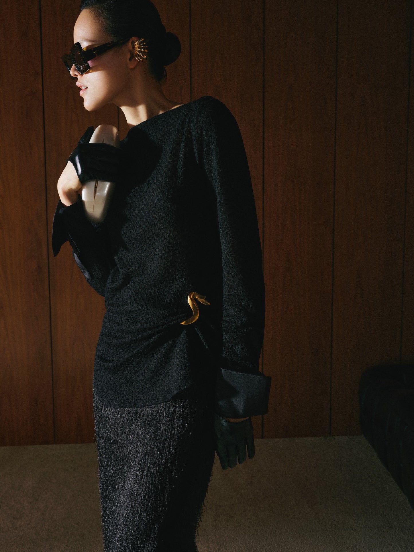 Woman wearing a black outfit with a gold belt against a wooden wall.