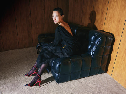 Woman in a black dress sitting on a black leather couch against a wooden paneled wall.