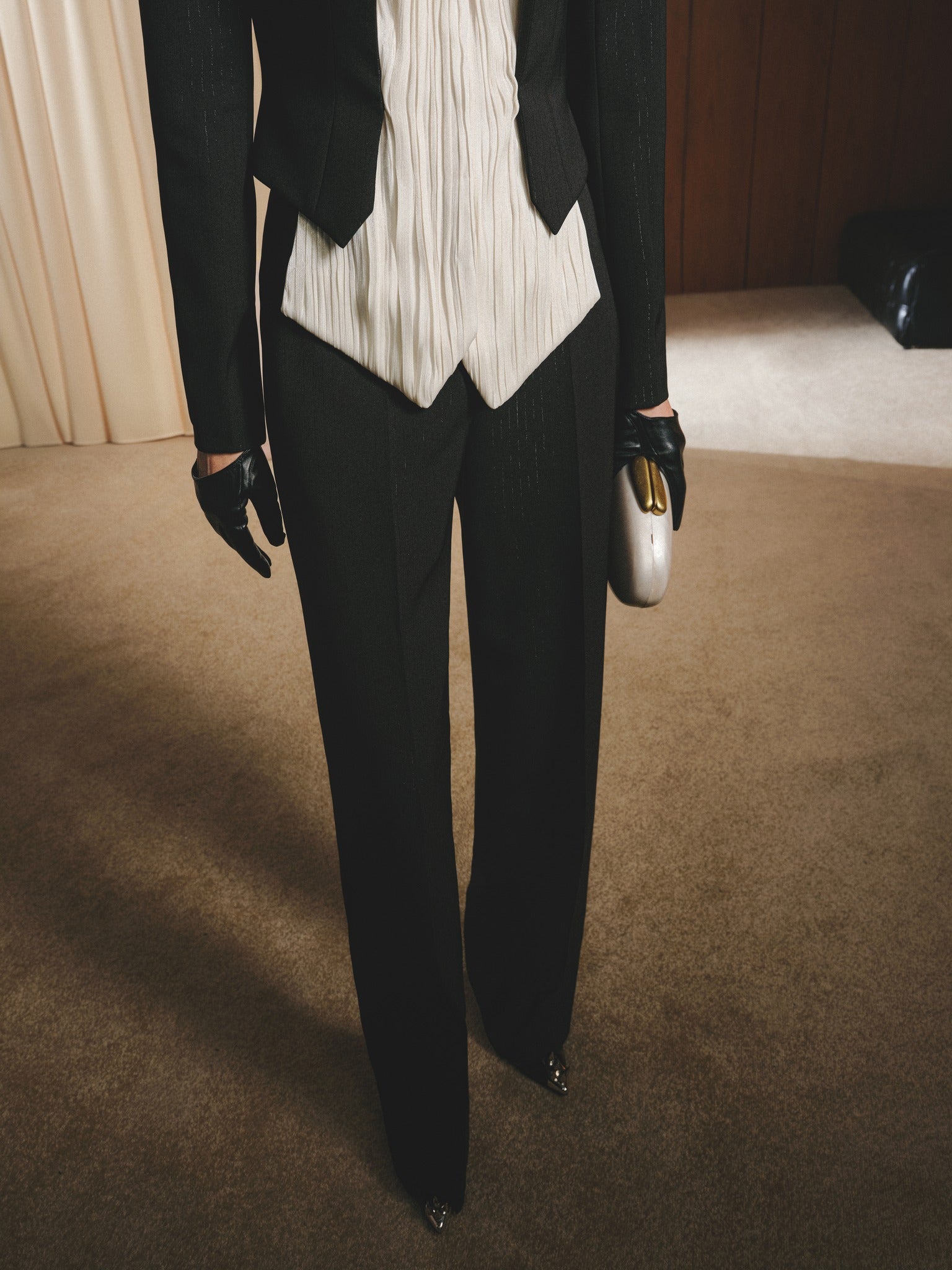 Person wearing a black blazer, white pleated shirt, and black pants on a brown carpet.