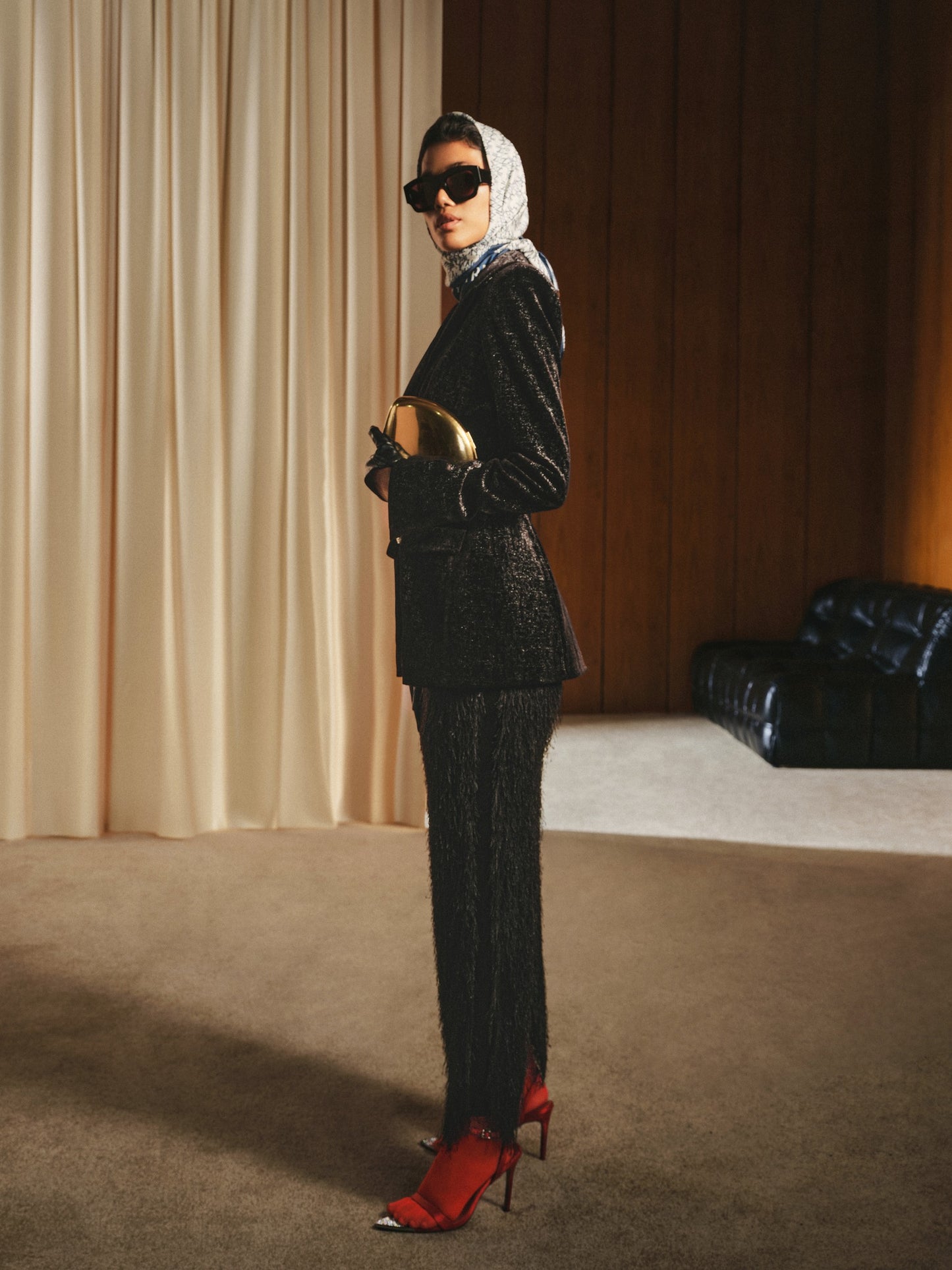 Woman in a black textured suit with red shoes standing in a room with beige curtains and a black chair.