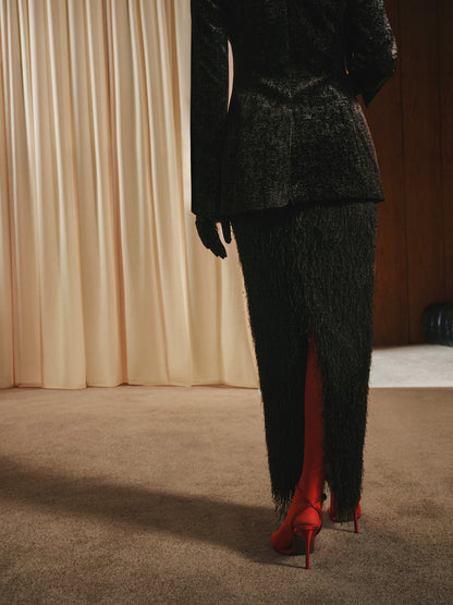 Person wearing a dark textured suit with red high heels in a room with beige curtains.