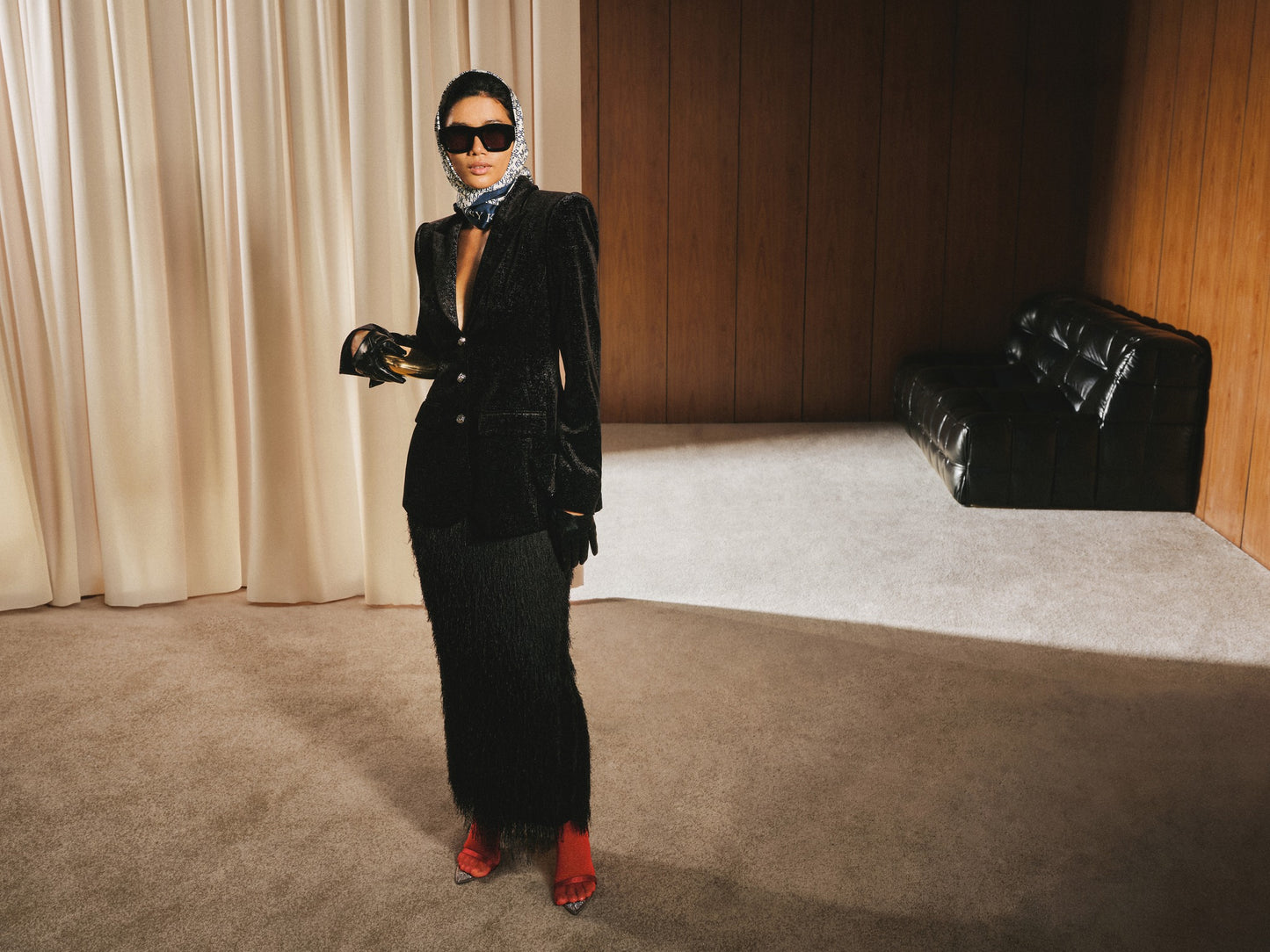 Woman in a black suit standing in a room with beige curtains and a black couch.