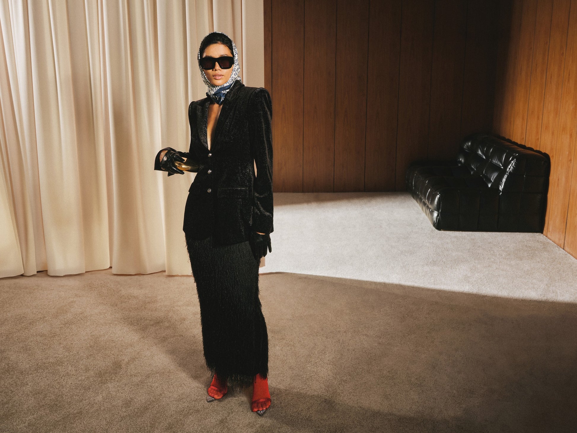 Woman in a black suit standing in a room with beige curtains and a black couch.