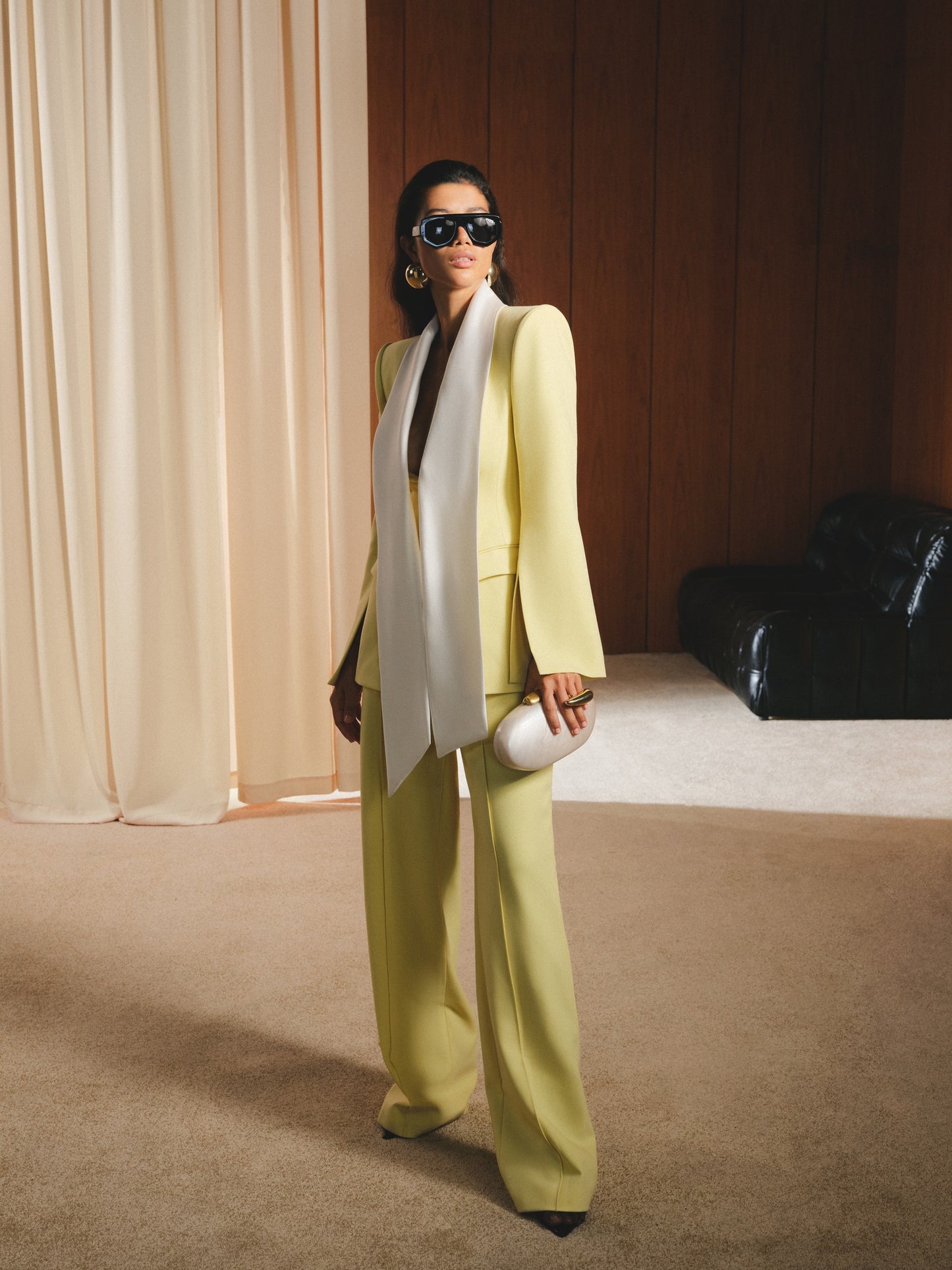 Woman in a light green suit with a white scarf standing in a room with beige curtains and a black bag.
