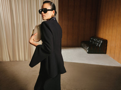 Person wearing a black outfit and sunglasses in a room with wooden walls and a black chair.