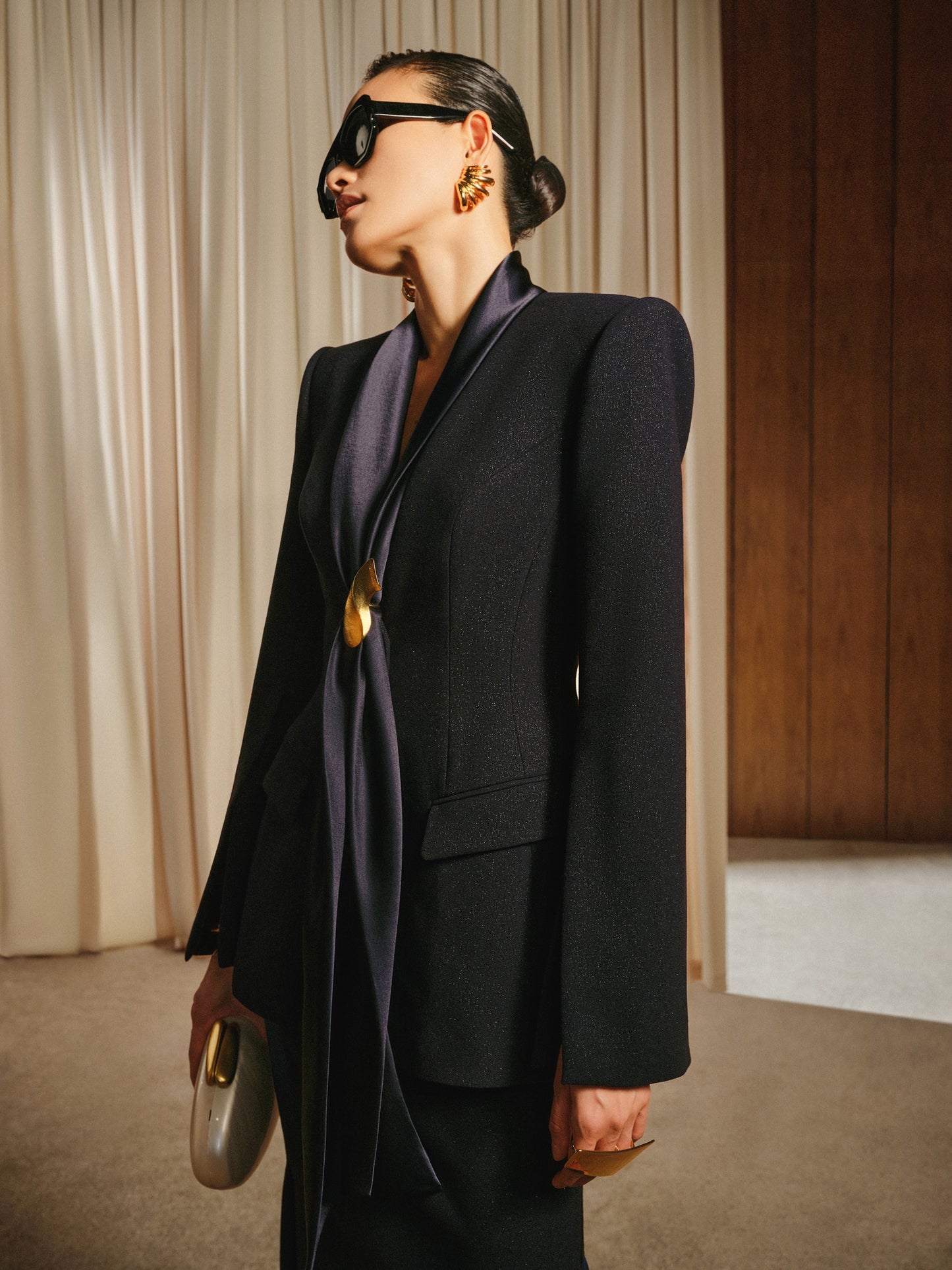 Person wearing a black suit with a long dark scarf, standing against a neutral background.