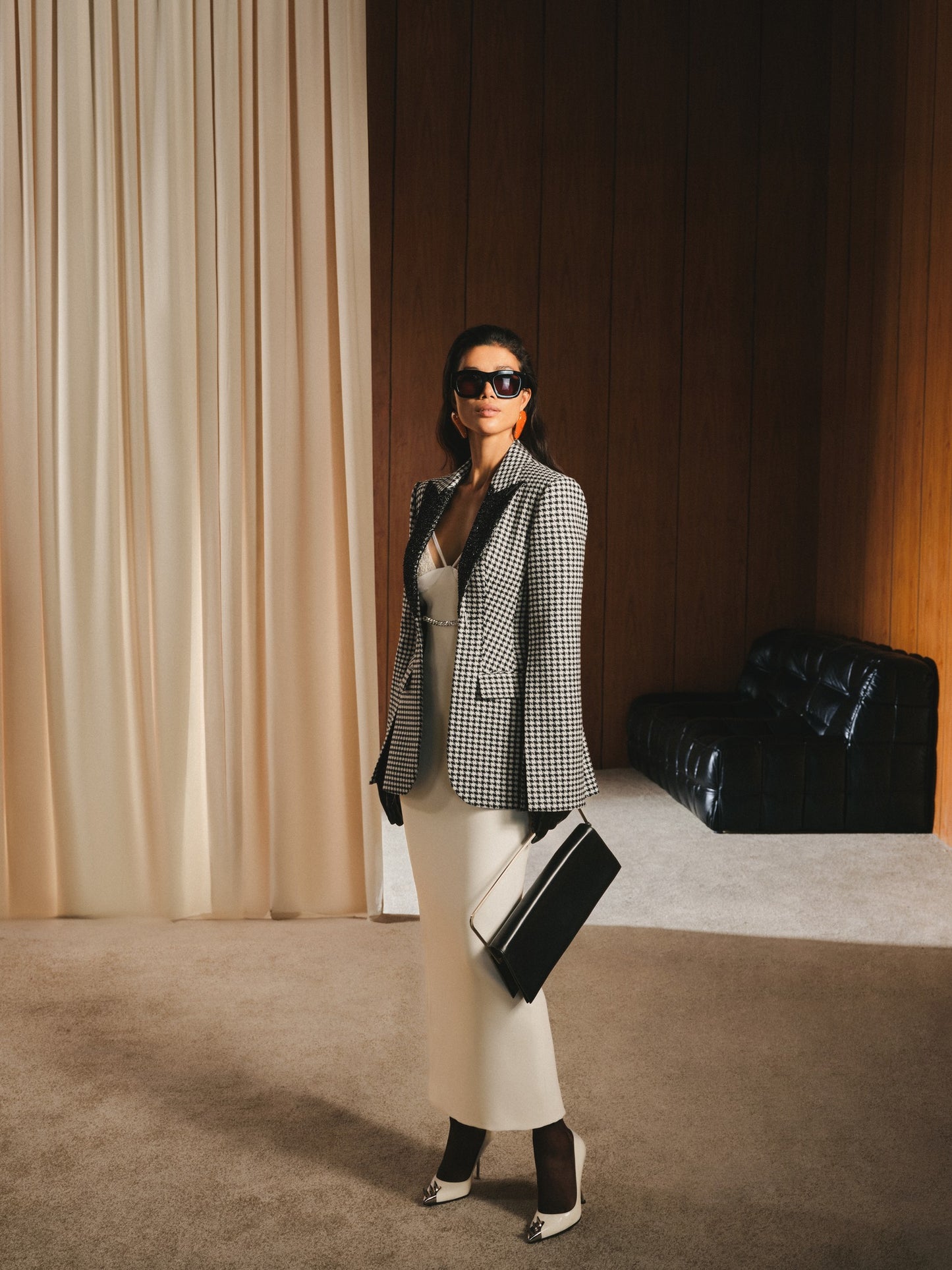 Woman in a checkered blazer and white pants standing in a room with curtains and a black chair.