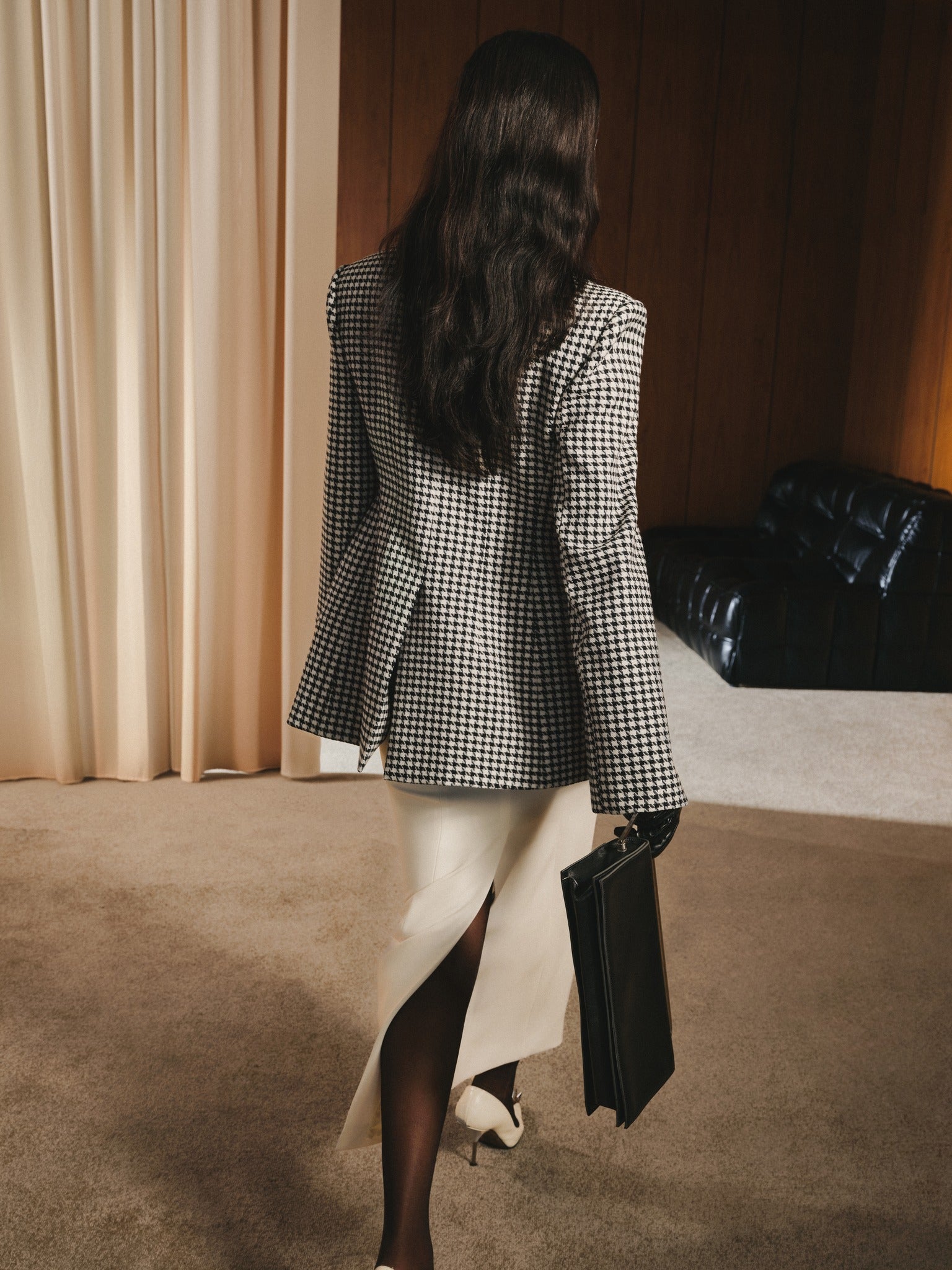 Woman in a checkered blazer and skirt holding a black clutch in an indoor setting.