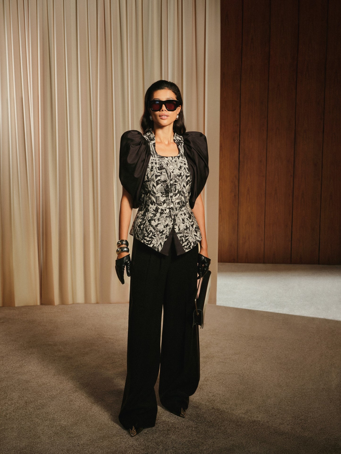 Woman in a stylish outfit with a floral top and black pants standing in a room with curtains and a wooden panel.