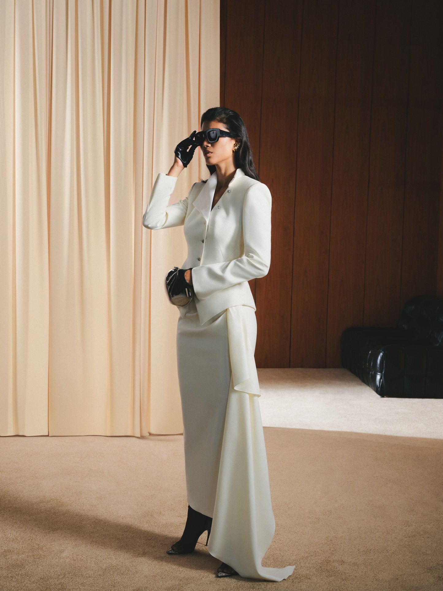 Woman in a white outfit standing in a room with beige curtains and wooden wall.
