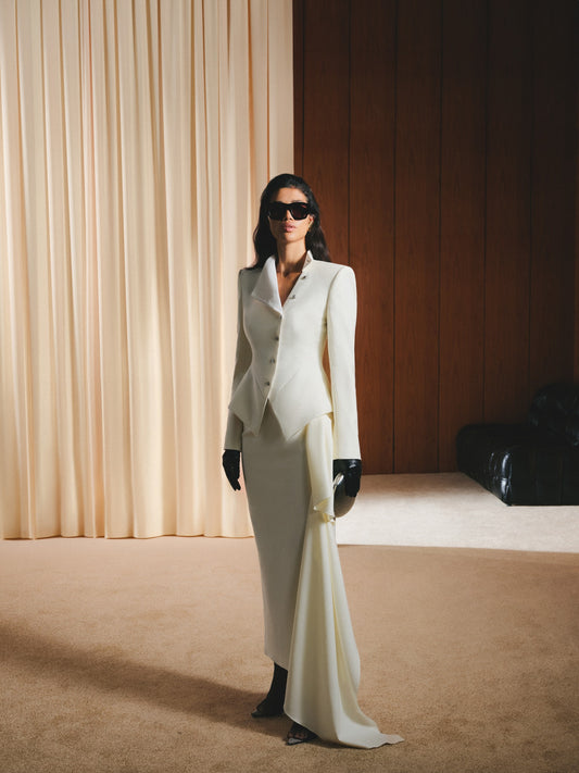 Woman in a white suit standing in a room with beige curtains and a black couch.