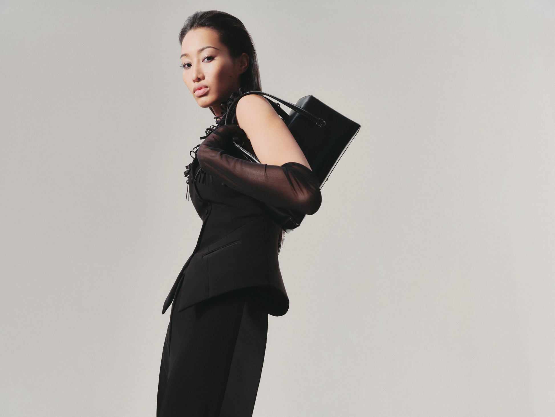 Woman in a black outfit holding a black handbag against a plain background