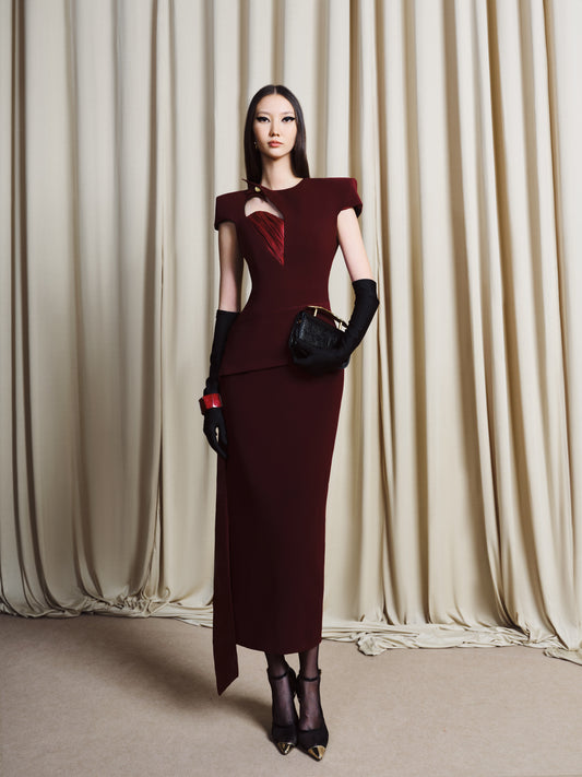 Woman in a burgundy evening gown with black gloves and shoes standing against a beige curtain.