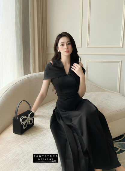 Woman in a black dress sitting on a couch with a handbag next to her, in a room with neutral decor.