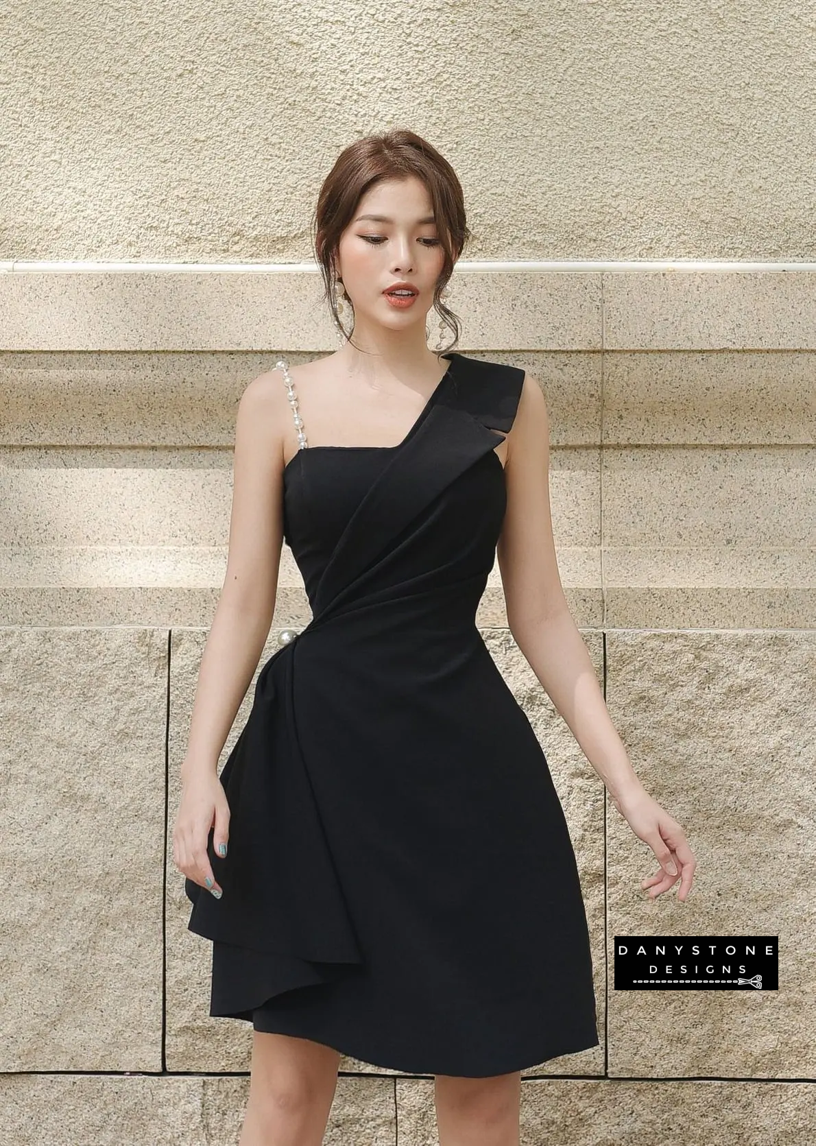 Model in Black One-Shoulder Pearl Strap Flared Dress Against Stone Wall