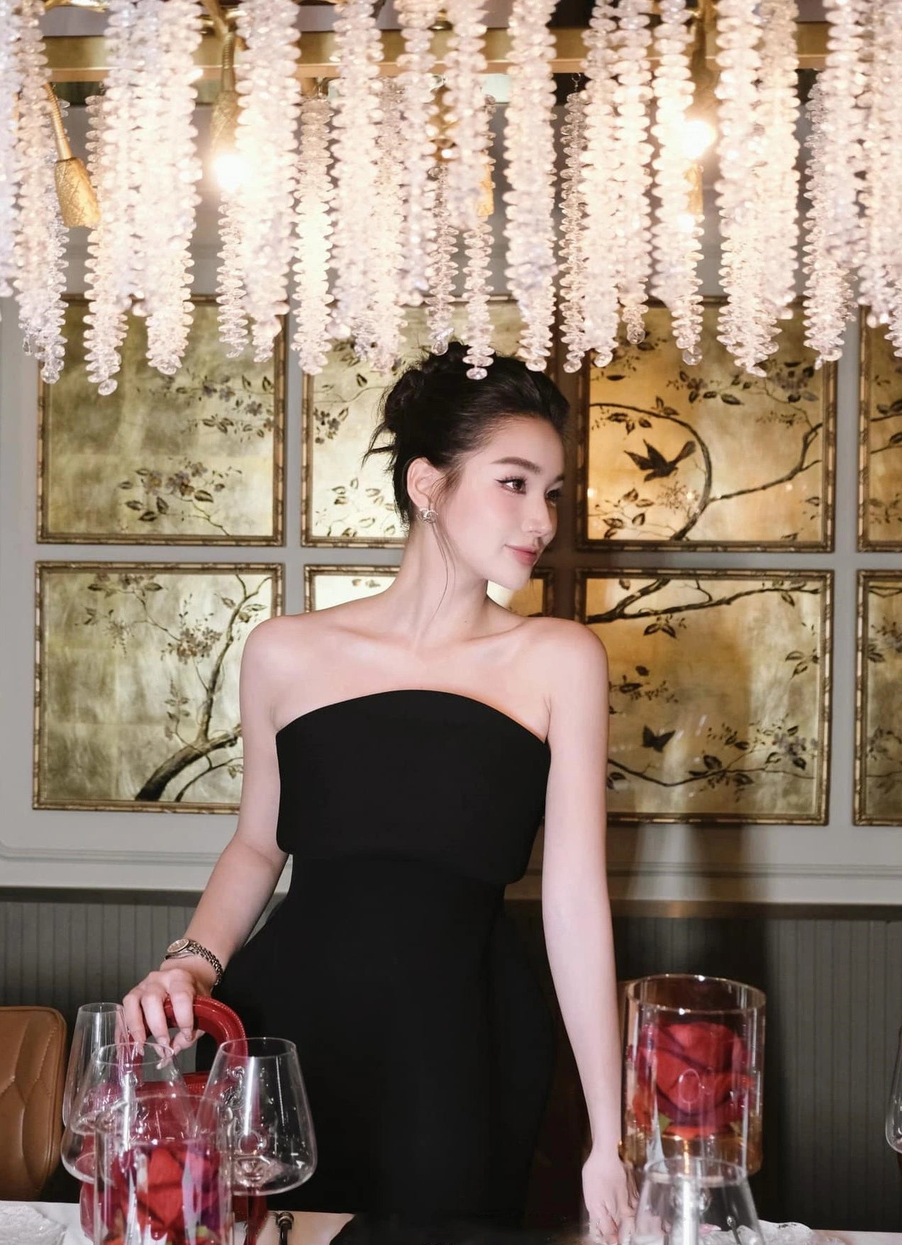 Woman in a black dress standing in a decorated room with chandelier and paintings.