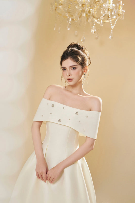 Woman wearing a white off-shoulder dress with embellishments against a soft yellow background.