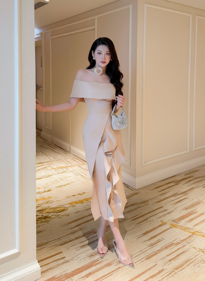 Woman in a beige off-shoulder dress standing in an elegant interior setting.
