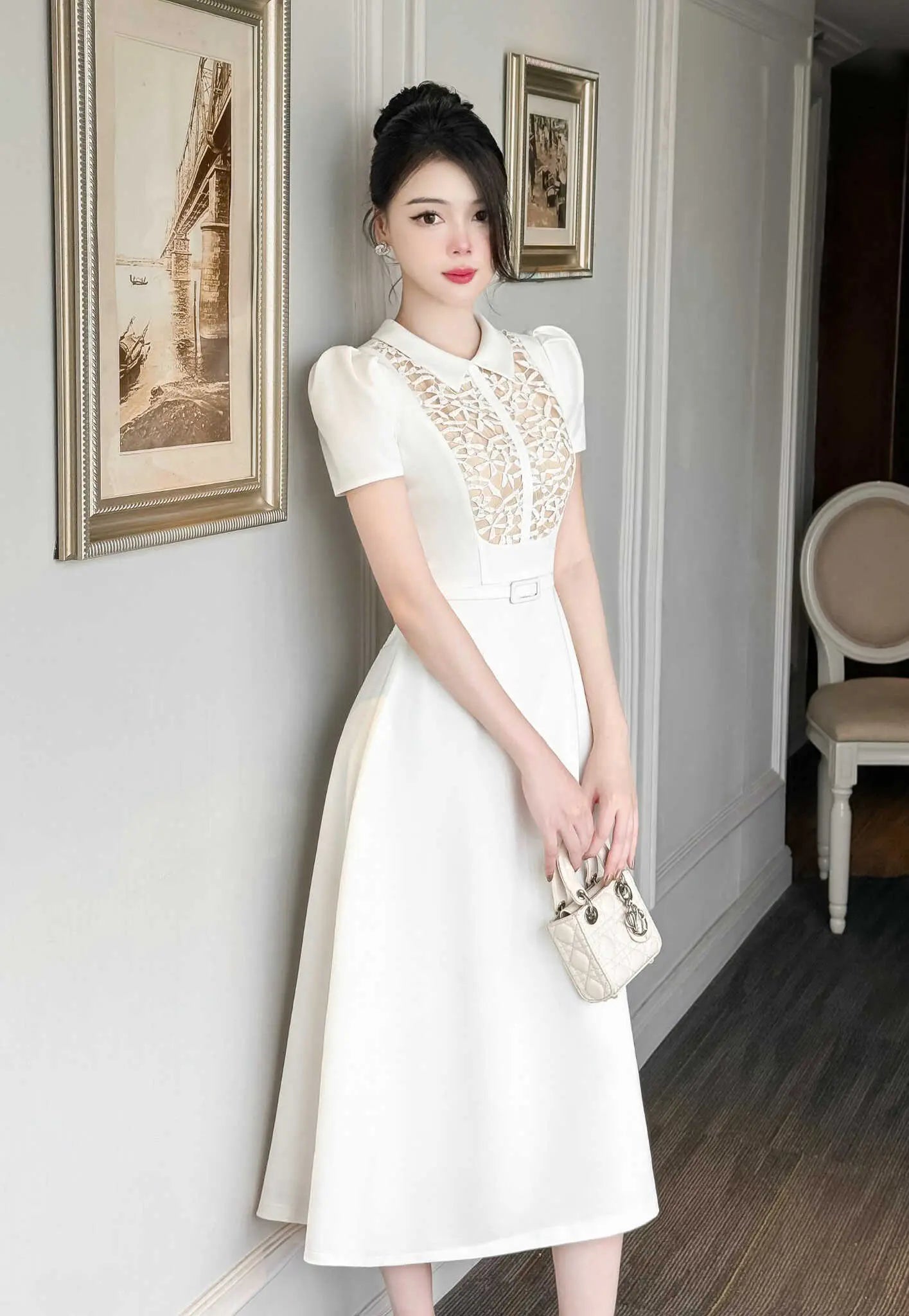 Woman in a white dress with lace details standing in a room with framed pictures on the wall.