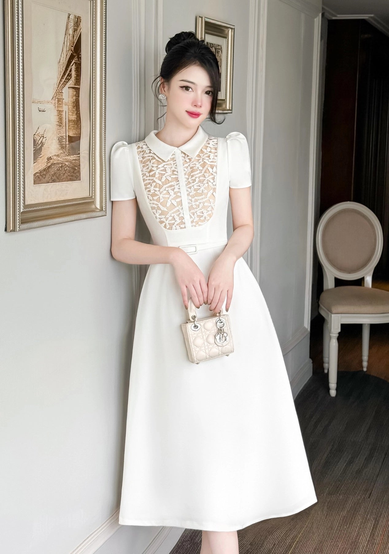 Woman in a white dress with lace details holding a handbag in an indoor setting.