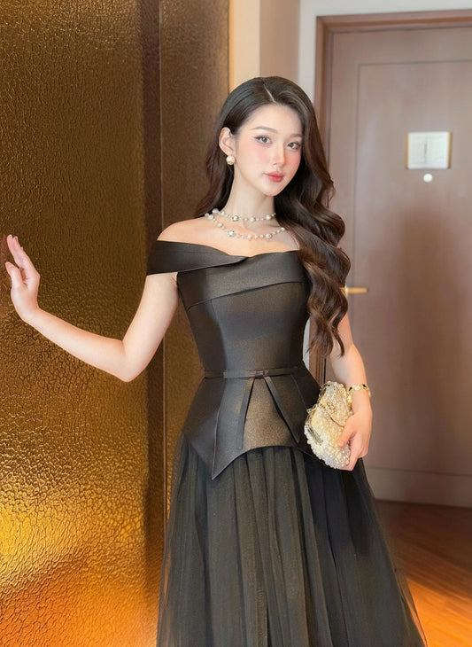 Woman in a black off-shoulder dress standing indoors with a textured wall and door in the background.