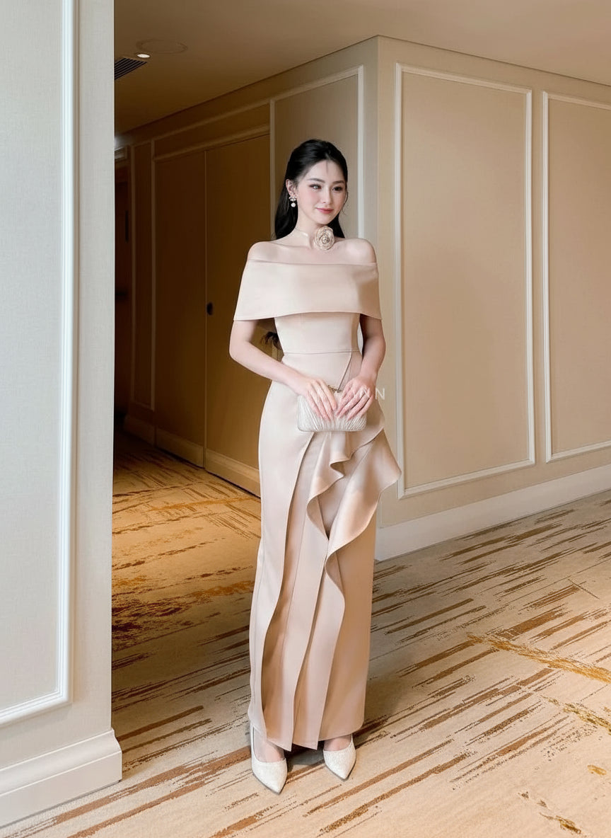 Woman in a beige off-shoulder dress standing in an elegant interior setting.