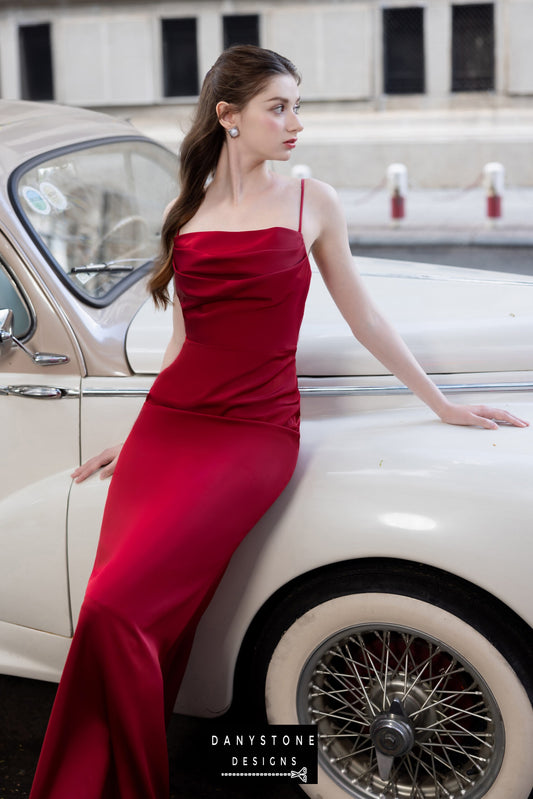Full-length red silk gown with open back and tie detail, worn by a model beside a vintage car.