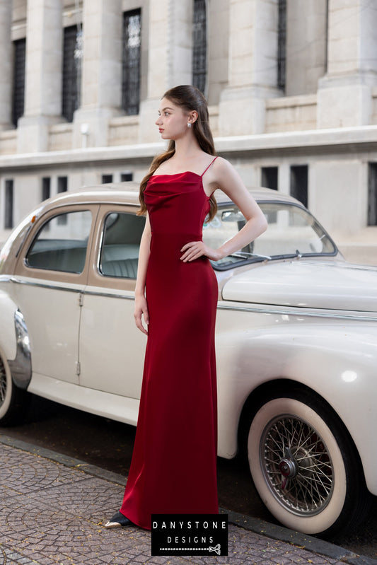 Model in a red silk evening gown with a cowl neckline and thin straps, standing beside a vintage car.