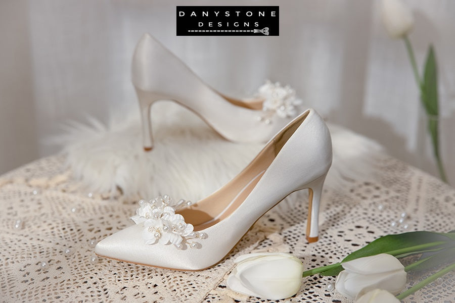 Elegant White Silk Satin Wedding Heels with floral design, one heel elevated in a glass vase, displayed on a lace tablecloth.