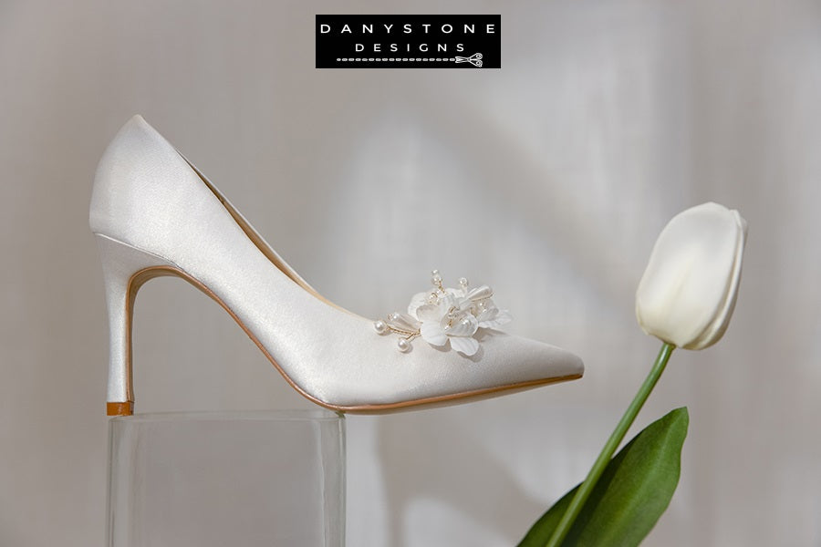 Close-up of Elegant White Silk Satin Wedding Heels with floral and pearl design on a lace tablecloth.