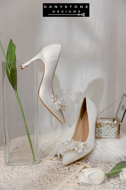 Pair of Elegant White Silk Satin Wedding Heels with floral embellishments, displayed with white tulips on a lace tablecloth.