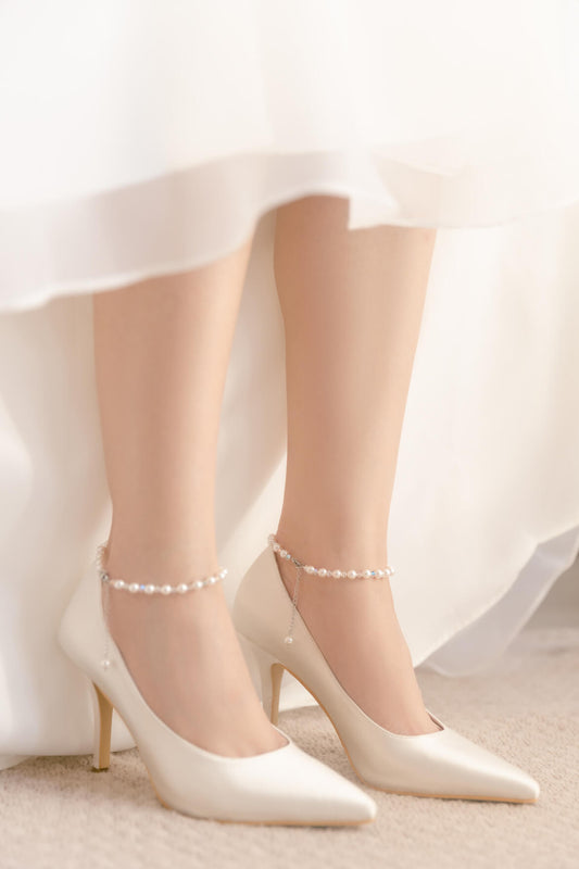 White high-heeled shoes with pearl ankle straps worn by a person in a white dress.