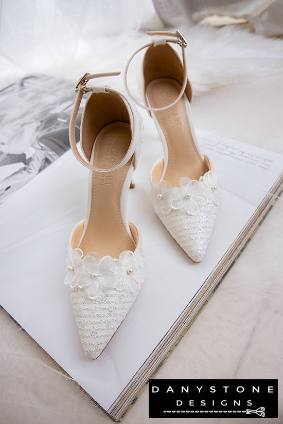 Elegant white lace bridal heels with ankle strap, 9cm heel, arranged with flowers in the background.