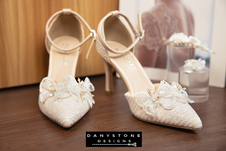 Top view of bridal shoes with chiffon butterfly wings and ankle straps, showcased on a wooden floor with a bridal dress in the background.