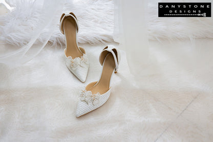 Top view of bridal shoes with chiffon butterfly wings and ankle straps, showcased on a wooden floor with a bridal dress in the background.